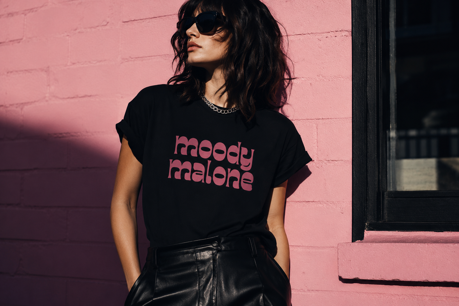 A woman wearing a black graphic T-shirt with 'moody malone' printed in pink retro lettering, paired with black leather pants, posing against a pink brick wall.